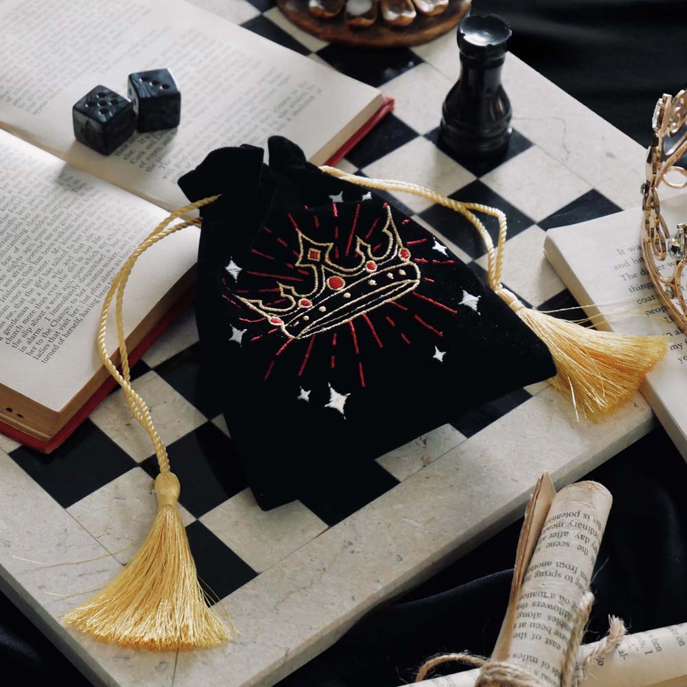 The Gilded Heir trinket pouch by The Quirky Cup Collective sits on a marble chess board on top of books. The black velvet pouch has gold drawstring and features a crown motif in gold, red and white embroidery.