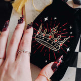 A black Quirky Cup Collective loot pouch features an intricate embroidered crown motif in red, gold and white. The pouch is held by a hand with intricate red nails and jewellery on a gaming table.