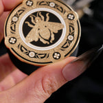 A hand with long dark nails is holding a The Quirky Cup Collective Clingie (it's like an enamel pop socket or phone grip!) in front of a black fabric background. This bookish inspired product featured a honey bee motif in gold surrounded by a circle design of fine flower elements.