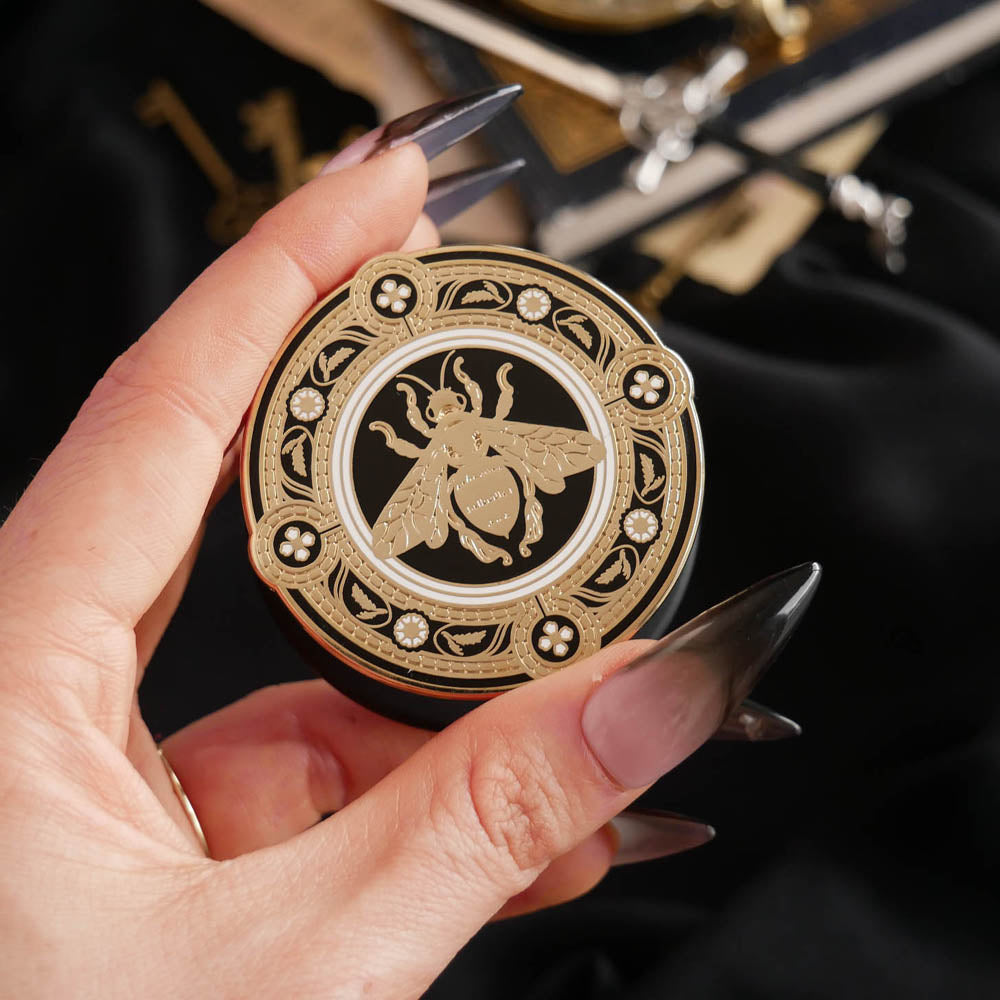 A hand with long dark nails is holding a The Quirky Cup Collective Clingie (it's like an enamel pop socket or phone grip!) in front of a black fabric background. This bookish inspired product featured a honey bee motif in gold surrounded by a circle design of fine flower elements.