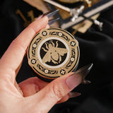 A hand with long dark nails is holding a The Quirky Cup Collective Clingie (it's like an enamel pop socket or phone grip!) in front of a black fabric background. This bookish inspired product featured a honey bee motif in gold surrounded by a circle design of fine flower elements.
