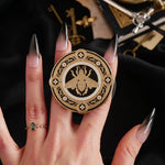 A hand with long dark nails is holding a The Quirky Cup Collective Clingie (it's like an enamel pop socket or phone grip!) in front of a black fabric background. This bookish inspired product featured a honey bee motif in gold surrounded by a circle design of fine flower elements.