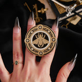 A hand with long dark nails is holding a The Quirky Cup Collective Clingie (it's like an enamel pop socket or phone grip!) in front of a black fabric background. This bookish inspired product featured a honey bee motif in gold surrounded by a circle design of fine flower elements.
