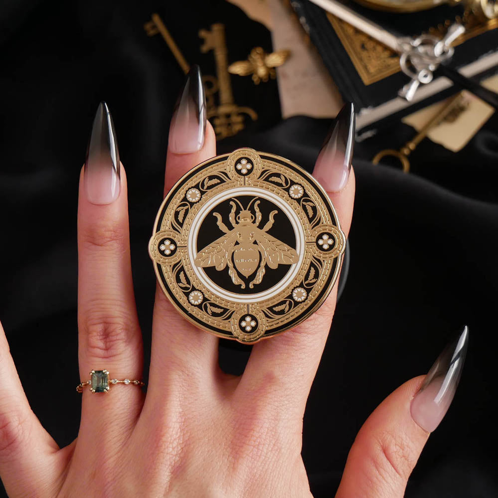 A hand with long dark nails is holding a The Quirky Cup Collective Clingie (it's like an enamel pop socket or phone grip!) in front of a black fabric background. This bookish inspired product featured a honey bee motif in gold surrounded by a circle design of fine flower elements.