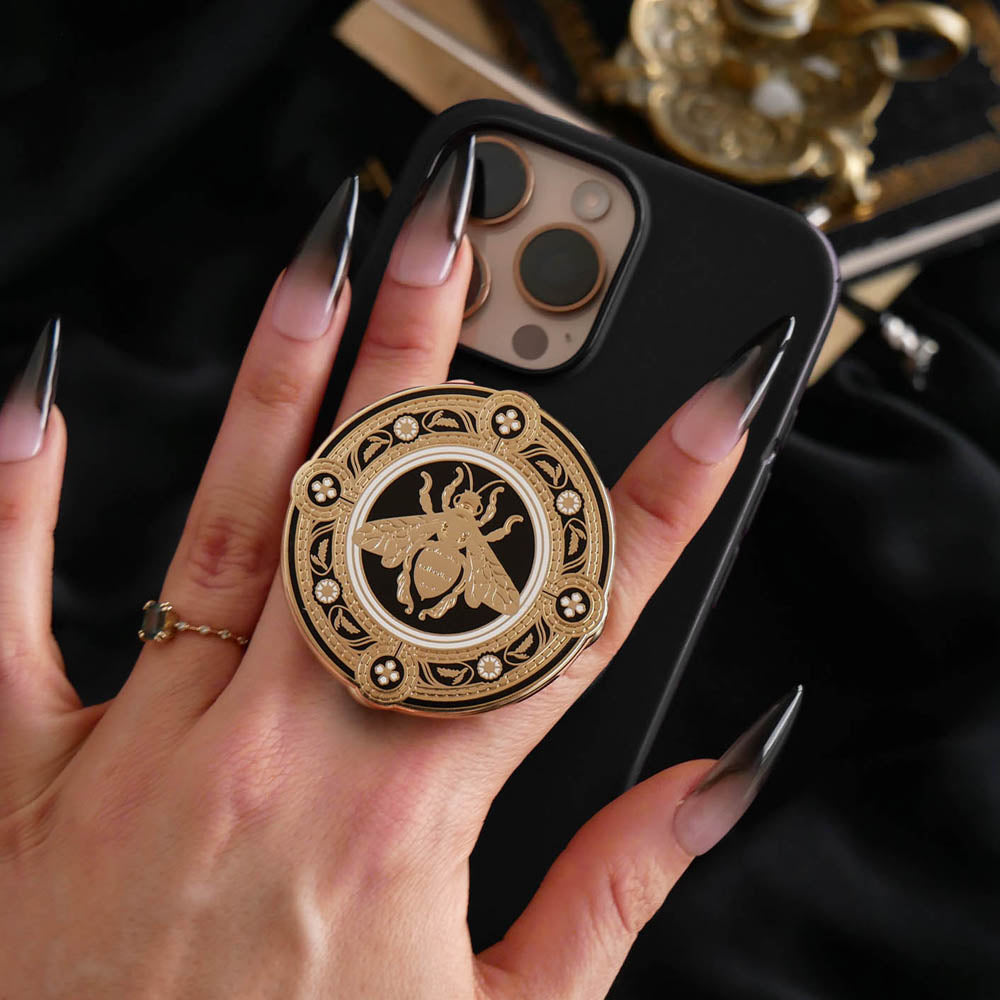 A hand with long dark nails is holding a The Quirky Cup Collective Clingie (it's like an enamel pop socket or phone grip!) on the back of an iPhone in front of a black fabric background. This bookish inspired product featured a honey bee motif in gold surrounded by a circle design of fine flower elements.