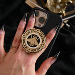 A hand with long dark nails is holding a The Quirky Cup Collective Clingie (it's like an enamel pop socket or phone grip!) on the back of an iPhone in front of a black fabric background. This bookish inspired product featured a honey bee motif in gold surrounded by a circle design of fine flower elements.