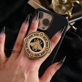 A hand with long dark nails is holding a The Quirky Cup Collective Clingie (it's like an enamel pop socket or phone grip!) on the back of an iPhone in front of a black fabric background. This bookish inspired product featured a honey bee motif in gold surrounded by a circle design of fine flower elements.
