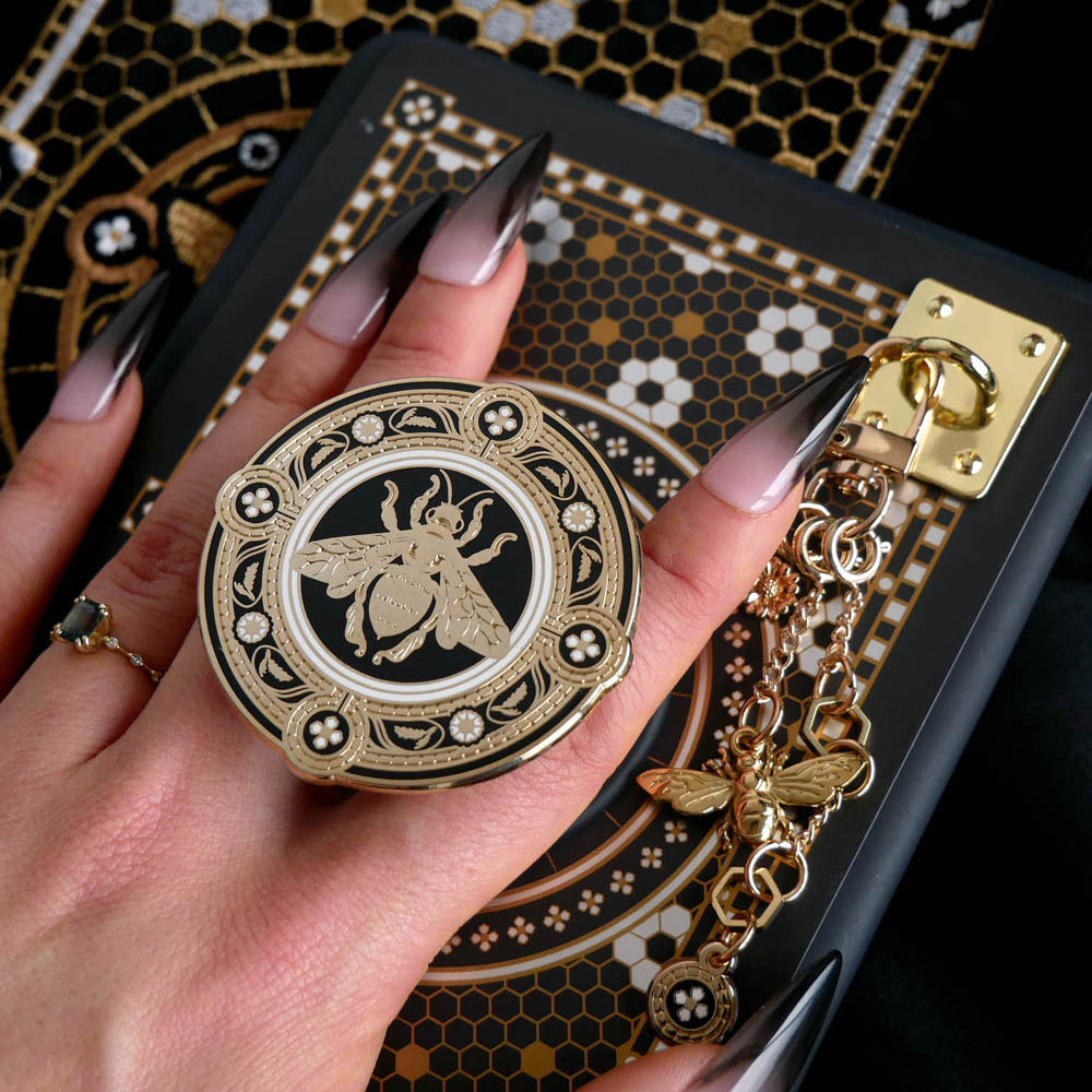 A hand with long dark nails is holding a The Quirky Cup Collective Clingie (it's like an enamel pop socket or phone grip!) in front of a black fabric background. This bookish inspired product featured a honey bee motif in gold surrounded by a circle design of fine flower elements.