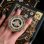 A hand with long dark nails is holding a The Quirky Cup Collective Clingie (it's like an enamel pop socket or phone grip!) in front of a black fabric background. This bookish inspired product featured a honey bee motif in gold surrounded by a circle design of fine flower elements.