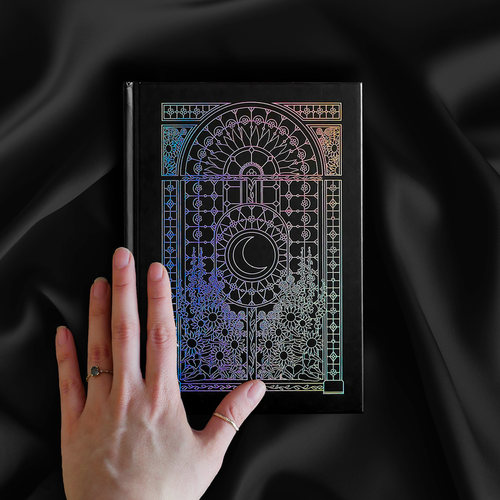 decorative black journal with holographic foiling sits on a background of black draped silk while the cover is being caressed by a hand