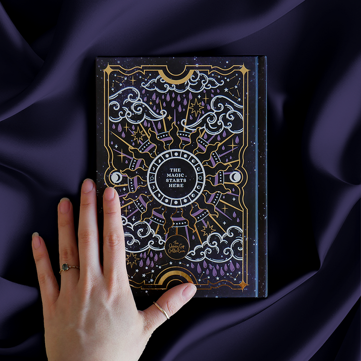 Journal with celestial cover sits on a purple silky background with a hand caressing the cover. Journal cover shows a moon nested inside a sun, surrounded by clouds, raindrops and stars. Foiling is purple and gold.