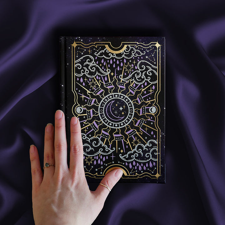 Journal with celestial cover sits on a purple silky background with a hand caressing the cover. Journal cover shows a moon nested inside a sun, surrounded by clouds, raindrops and stars. Foiling is purple and gold.