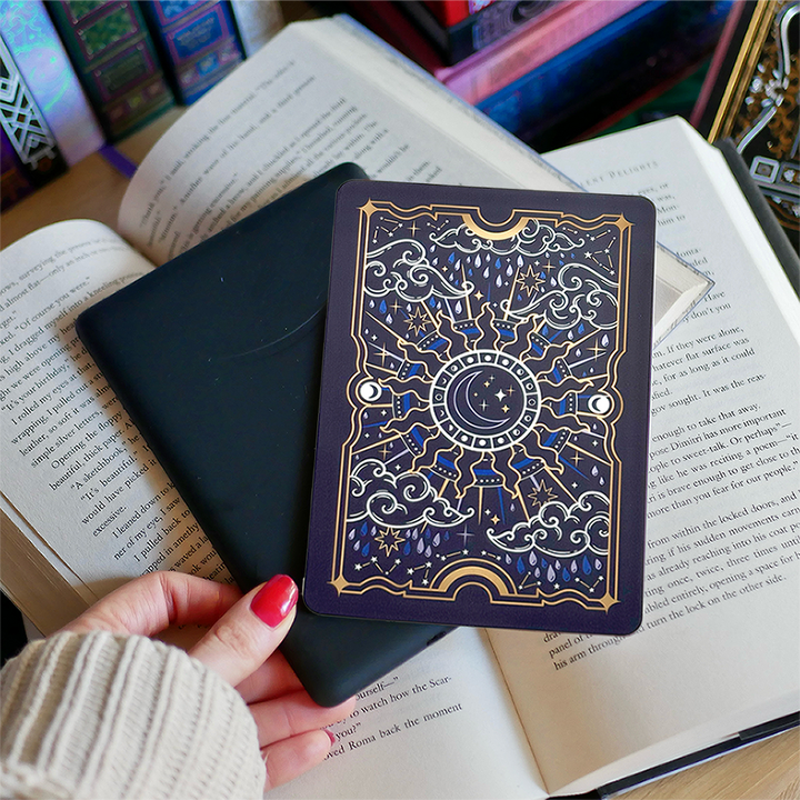 Image shows a hand gently touching a kindle and a kindle sticker insert sitting on top of the device. They sit on a bed of open books. The navy, blue, gold  and white design shows a moon in the centre, surrounded by sun rays, clouds, stars, raindrops and decorative celestial embellishments. 