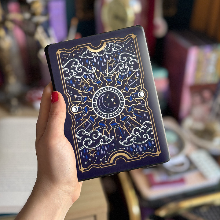 Image shows a hand holding a kindle showing off a kindle sticker insert that is placed on the back of the device. The navy, blue, gold  and white design shows a moon in the centre, surrounded by sun rays, clouds, stars, raindrops and decorative celestial embellishments. 