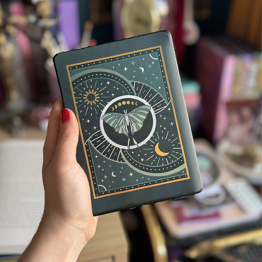 Image shows a hand holding a kindle showing off a kindle sticker insert that is placed on the back of the device. The teal, gold and white design shows a luna moth surrounded by moons, stars and decorative embellishments. 