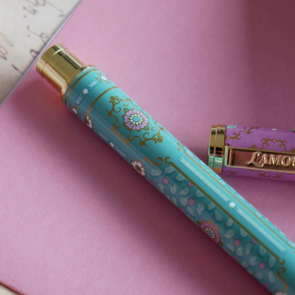 The L'amour L'emporte pen sits in front of a feminine french journaling set up. The french inspired gel pen features floral motifs in pink and aqua inspired by a classic literature aesthetic and 'L'AMOUR L'EMPORTE' etched into the pen clip.