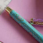 The L'amour L'emporte pen sits in front of a feminine french journaling set up. The french inspired gel pen features floral motifs in pink and aqua inspired by a classic literature aesthetic and 'L'AMOUR L'EMPORTE' etched into the pen clip.