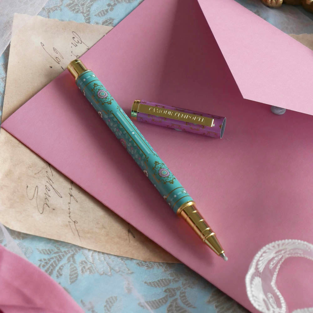 The L'amour L'emporte pen sits on a french journaling set up. The french inspired gel pen features floral motifs in pink and aqua inspired by a classic literature aesthetic and 'L'AMOUR L'EMPORTE' etched into the pen clip.