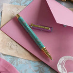 The L'amour L'emporte pen sits on a french journaling set up. The french inspired gel pen features floral motifs in pink and aqua inspired by a classic literature aesthetic and 'L'AMOUR L'EMPORTE' etched into the pen clip.