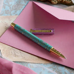 The L'amour L'emporte pen sits on a pink background in a feminine journaling aesthetic. The french inspired gel pen features floral motifs in pink and aqua inspired by a classic literature aesthetic and 'L'AMOUR L'EMPORTE' etched into the pen clip.