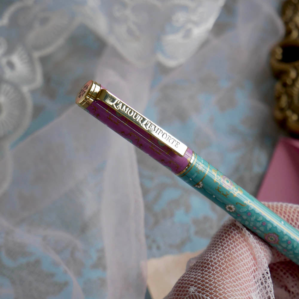 The L'amour L'emporte pen is held in a lace gloved hand in front of a feminine french journaling set up. The french inspired gel pen features floral motifs in pink and aqua inspired by a classic literature aesthetic and 'L'AMOUR L'EMPORTE' etched into the pen clip.