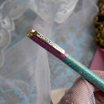 The L'amour L'emporte pen is held in a lace gloved hand in front of a feminine french journaling set up. The french inspired gel pen features floral motifs in pink and aqua inspired by a classic literature aesthetic and 'L'AMOUR L'EMPORTE' etched into the pen clip.