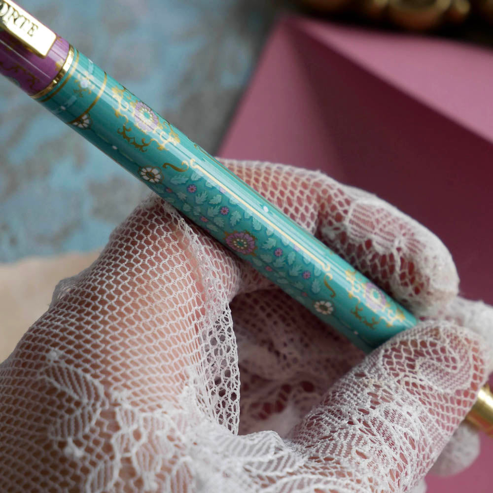 The L'amour L'emporte pen is held in a lace gloved hand in front of a feminine french journaling set up. The french inspired gel pen features floral motifs in pink and aqua inspired by a classic literature aesthetic and 'L'AMOUR L'EMPORTE' etched into the pen clip.