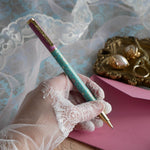 The L'amour L'emporte pen is held in a lace gloved hand in front of a feminine french journaling set up. The french inspired gel pen features floral motifs in pink and aqua inspired by a classic literature aesthetic and 'L'AMOUR L'EMPORTE' etched into the pen clip.