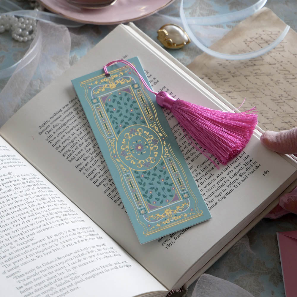 The ornate Antoinette light blue bookmark with gold detailing and a bright pink tassel rests on an open book. The light blue foiled bookmark featured gold, pink, cream and green foiling for a  Victorian inspired reading aesthetic.