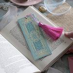 The ornate Antoinette light blue bookmark with gold detailing and a bright pink tassel rests on an open book. The light blue foiled bookmark featured gold, pink, cream and green foiling for a  Victorian inspired reading aesthetic.