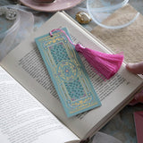 The ornate Antoinette light blue bookmark with gold detailing and a bright pink tassel rests on an open book. The light blue foiled bookmark featured gold, pink, cream and green foiling for a  Victorian inspired reading aesthetic.