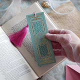 The ornate Antoinette light blue bookmark with gold detailing and a bright pink tassel rests on an open book. The light blue foiled bookmark featured gold, pink, cream and green foiling for a  Victorian inspired reading aesthetic.