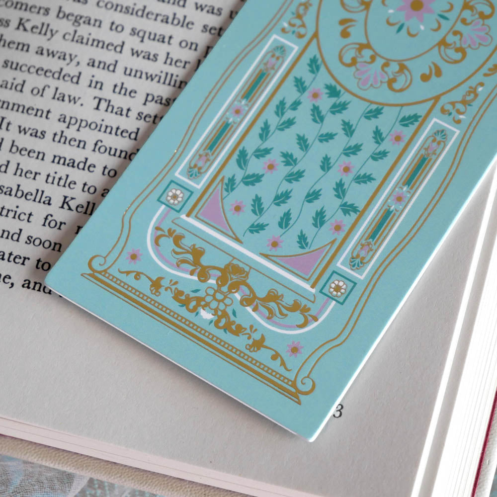 A detail of the ornate Antoinette light blue bookmark with gold detailing and a bright pink tassel rests on an open book. The light blue foiled bookmark featured gold, pink, cream and green foiling for a  classic book inspired reading aesthetic.