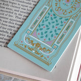 A detail of the ornate Antoinette light blue bookmark with gold detailing and a bright pink tassel rests on an open book. The light blue foiled bookmark featured gold, pink, cream and green foiling for a  classic book inspired reading aesthetic.
