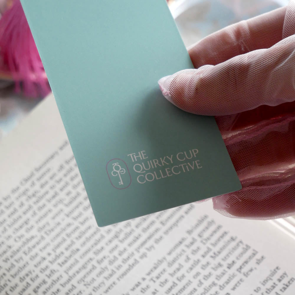 A detail of the back of the Antoinette light blue bookmark with gold detailing and a bright pink tassel rests on an open book. The light blue foiled bookmark is the perfect addition to the classic book inspired reading aesthetic.