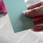A detail of the back of the Antoinette light blue bookmark with gold detailing and a bright pink tassel rests on an open book. The light blue foiled bookmark is the perfect addition to the classic book inspired reading aesthetic.