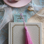 A detail of the back of the Antoinette light blue bookmark with gold detailing and a bright pink tassel sticks out the front of a white classic book. The light blue foiled bookmark is the perfect addition to the victorian bookish inspired reading aesthetic.