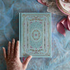The Antoinette hardcover notebook features an ornate pastel cover design, and sits on a blue lace fabric surface. The french aesthetic journal features intricate gold, pink, cream and aqua foiling with a design inspired by classical literature and academia reading aesthetic.