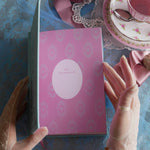 The Antoinette foiled hardcover journal sits open with he pink title pages showing a nameplate to be filled out. The scene is styled with a pink teacup and saucer, pearls, and a blue patterned fabric background for a french inspired journal aesthetic.