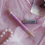 Bibliophile pen in the pastel pink and aqua Antoinette colour-way sits on a pink lace background surrounded by pearls. The french inspired gel pen features floral motifs inspired by a classic literature aesthetic and 'BIBLIOPHILE' etched into the pen clip.