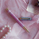 Bibliophile pen in the pastel pink and aqua Antoinette colour-way sits on a pink lace background surrounded by pearls. The french inspired gel pen features floral motifs inspired by a classic literature aesthetic and 'BIBLIOPHILE' etched into the pen clip.
