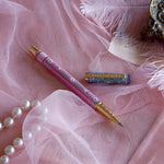 Bibliophile pen in the pastel pink and aqua Antoinette colour-way sits on a pink lace background surrounded by pearls. The french inspired gel pen features floral motifs inspired by a classic literature aesthetic and 'BIBLIOPHILE' etched into the pen clip.