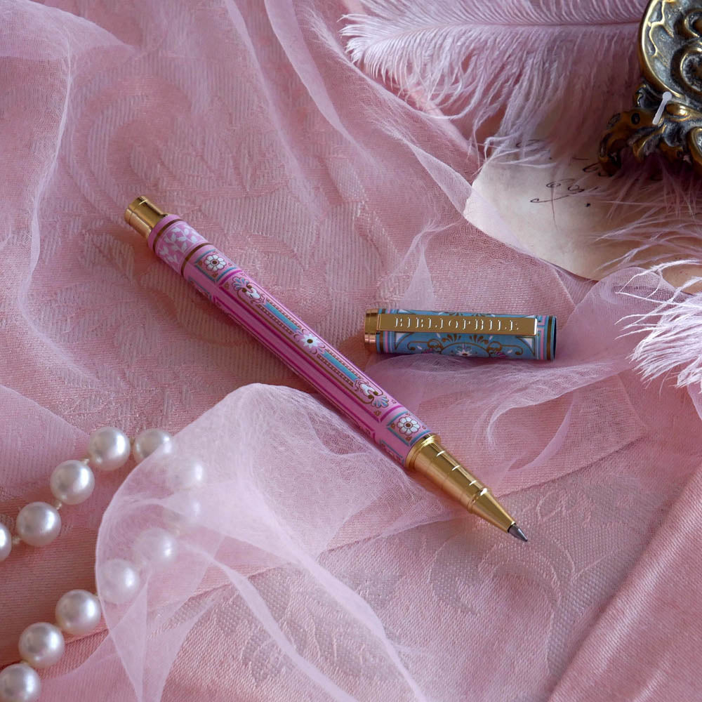 Bibliophile pen in the pastel pink and aqua Antoinette colour-way sits on a pink lace background surrounded by pearls. The french inspired gel pen features floral motifs inspired by a classic literature aesthetic and 'BIBLIOPHILE' etched into the pen clip.