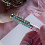 A detail of the Bibliophile pen in the pastel pink and aqua Antoinette colour-way sits on a pink lace background surrounded by pearls. The french inspired gel pen features floral motifs inspired by a classic literature aesthetic and 'BIBLIOPHILE' etched into the pen clip.