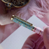 A detail of the Bibliophile pen in the pastel pink and aqua Antoinette colour-way sits on a pink lace background surrounded by pearls. The french inspired gel pen features floral motifs inspired by a classic literature aesthetic and 'BIBLIOPHILE' etched into the pen clip.