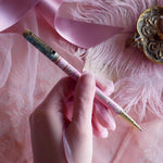 Bibliophile pen in the pastel pink and aqua Antoinette colour-way is held in a lace glove hand over a pink fabric background. The french inspired gold gel pen features floral motifs inspired by a classic literature aesthetic and 'BIBLIOPHILE' etched into the pen clip.