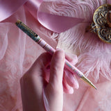 Bibliophile pen in the pastel pink and aqua Antoinette colour-way is held in a lace glove hand over a pink fabric background. The french inspired gold gel pen features floral motifs inspired by a classic literature aesthetic and 'BIBLIOPHILE' etched into the pen clip.