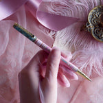 Bibliophile pen in the pastel pink and aqua Antoinette colour-way is held in a lace glove hand over a pink fabric background. The french inspired gel pen features floral motifs inspired by a classic literature aesthetic and 'BIBLIOPHILE' etched into the pen clip.