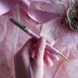 Bibliophile pen in the pastel pink and aqua Antoinette colour-way is held in a lace glove hand over a pink fabric background. The french inspired gel pen features floral motifs inspired by a classic literature aesthetic and 'BIBLIOPHILE' etched into the pen clip.