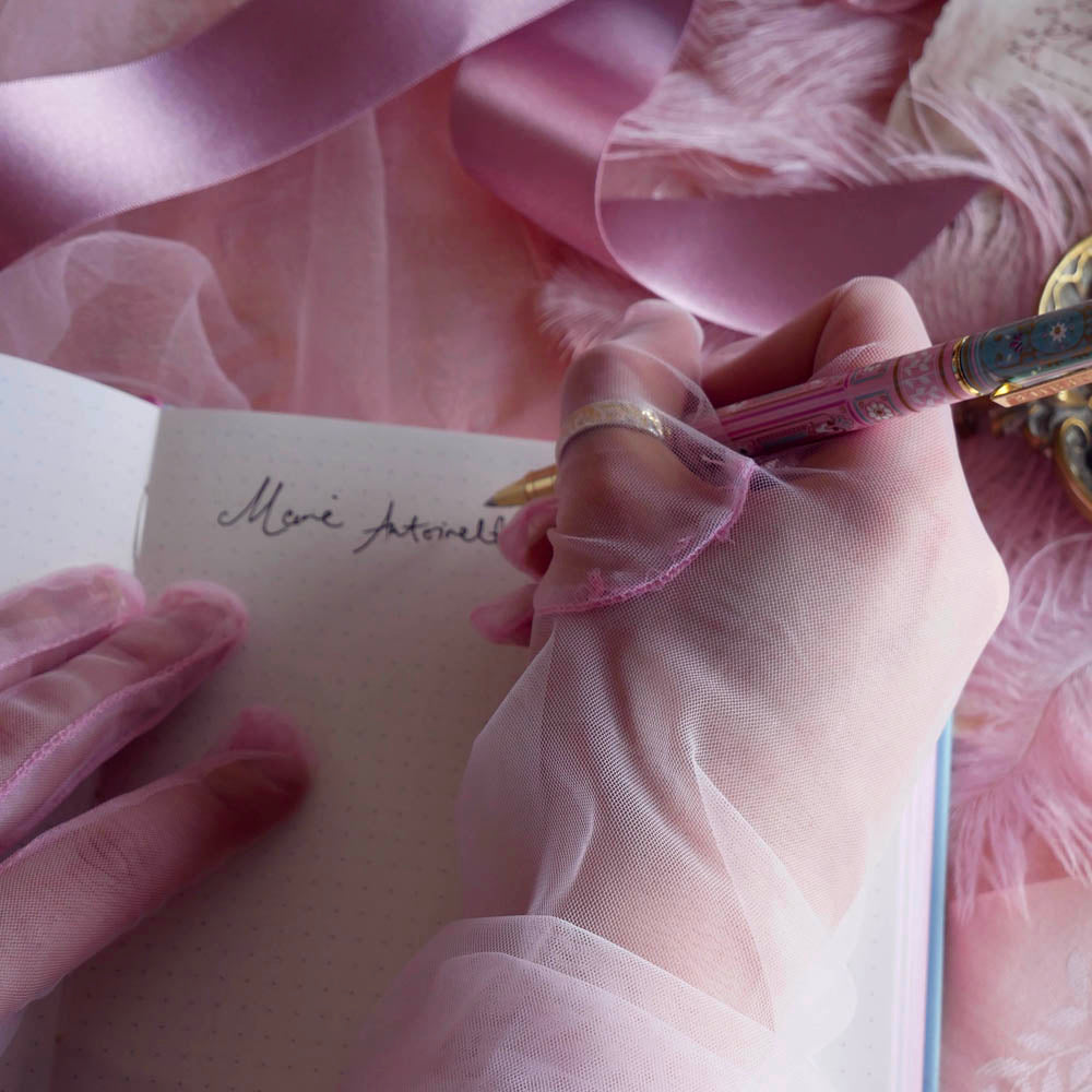 Bibliophile pen in the pastel pink and aqua Antoinette colour-way is held in a lace glove hand as it writes in the matching dot grid journal. The french inspired gel pen features floral motifs inspired by a classic literature aesthetic and 'BIBLIOPHILE' etched into the pen clip.